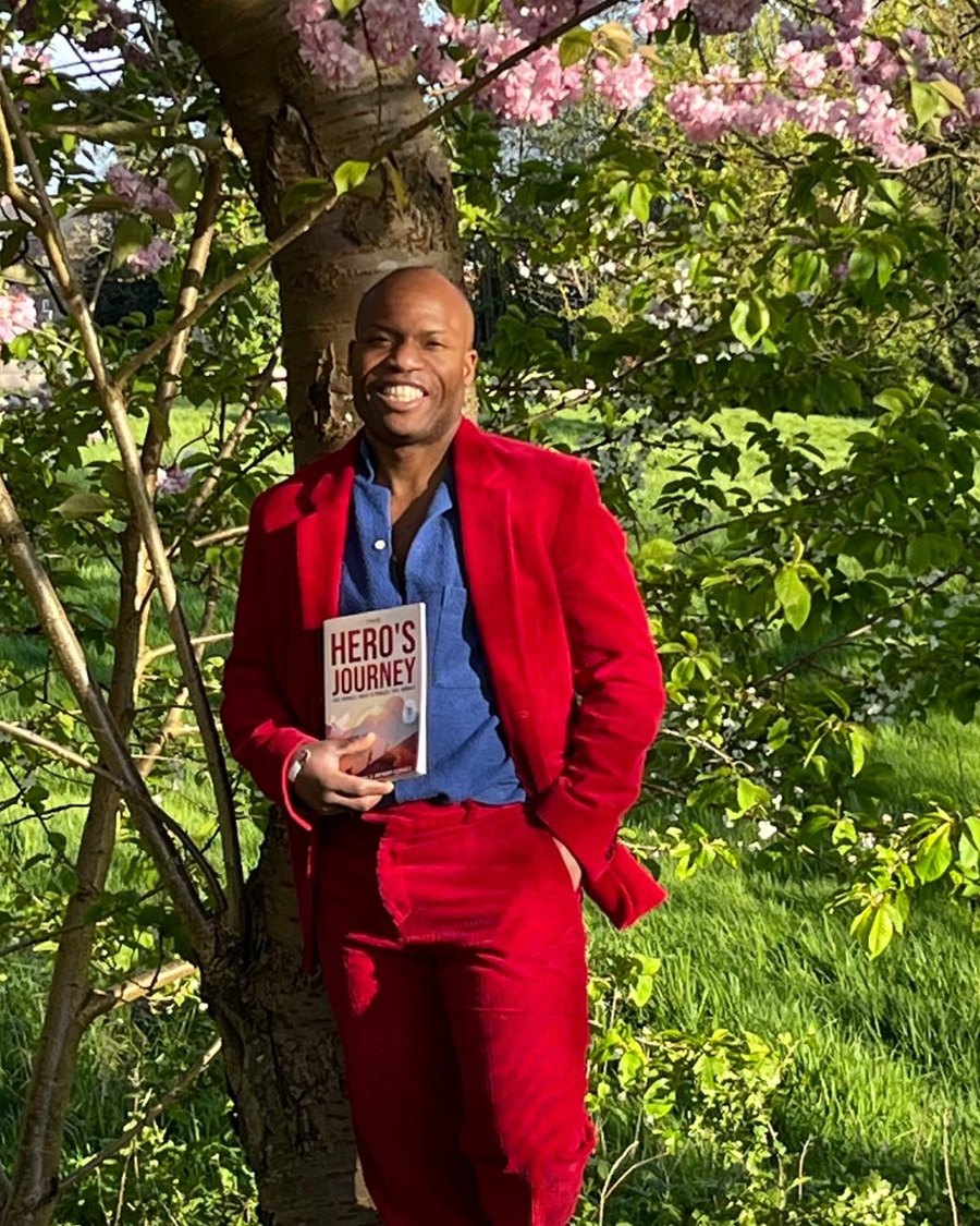 Nelson Lecuane standing beneath blossom in a red suit, holding a copy of his book The Hero's Journey